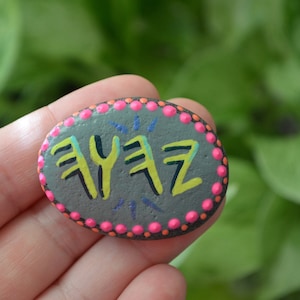 May include: A grey painted rock with a pink and orange dotted border. The rock has the word "YFZ" painted in yellow and green.