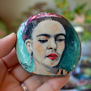 May include: Hand-painted rock art featuring a portrait of Frida Kahlo with a colourful floral headband, turquoise background, and the artist's signature "Hiso" in the bottom left corner.