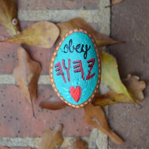 May include: A painted rock with a turquoise background, pink polka dots, and the words "obey" and "YEEZ" in black. A red heart is painted in the centre.
