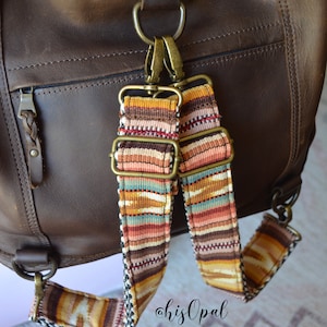 May include: A brown leather bag with a woven strap. The strap is attached to the bag with a brass buckle. The bag has a zipper closure and a small pocket on the front. The bag is sitting on a white surface. #hisOpal