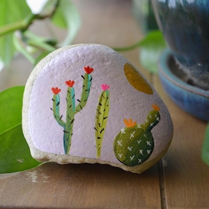 May include: Hand-painted rock with a purple background featuring a cactus with pink and red flowers and a prickly pear cactus with yellow flowers.