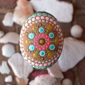May include: A round, brown stone painted with a colourful mandala design. The mandala features turquoise, pink, white, gold, green, and orange dots.
