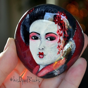 May include: A hand-painted rock depicting a geisha with red and white flowers in her hair. The background is a deep red colour. The rock is painted in a realistic style. #hisOpalRocks