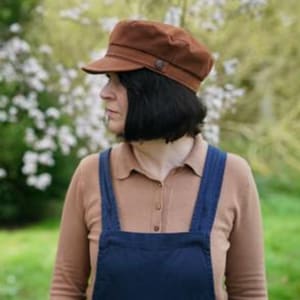 May include: A brown corduroy cap with a slightly curved brim. The cap has a button on the top and is being worn by a person standing in a garden.