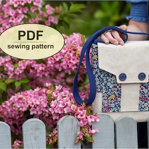 Peut inclure: Un patron de couture PDF pour un petit sac bandoulière avec une fermeture à rabat et deux boutons bleus. Le sac est fait d'un tissu en lin beige avec une doublure à imprimé floral. Le sac est présenté devant un buisson de fleurs roses et une clôture en bois blanc.