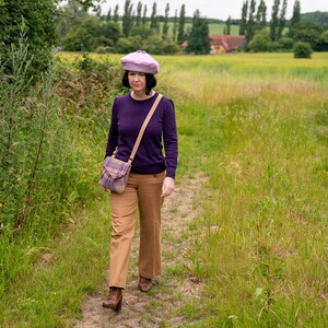 Pu&ograve; includere: Una donna con un maglione viola, pantaloni marroni e un berretto viola chiaro cammina su un sentiero di terra in un campo. Porta una borsa a tracolla a quadri.