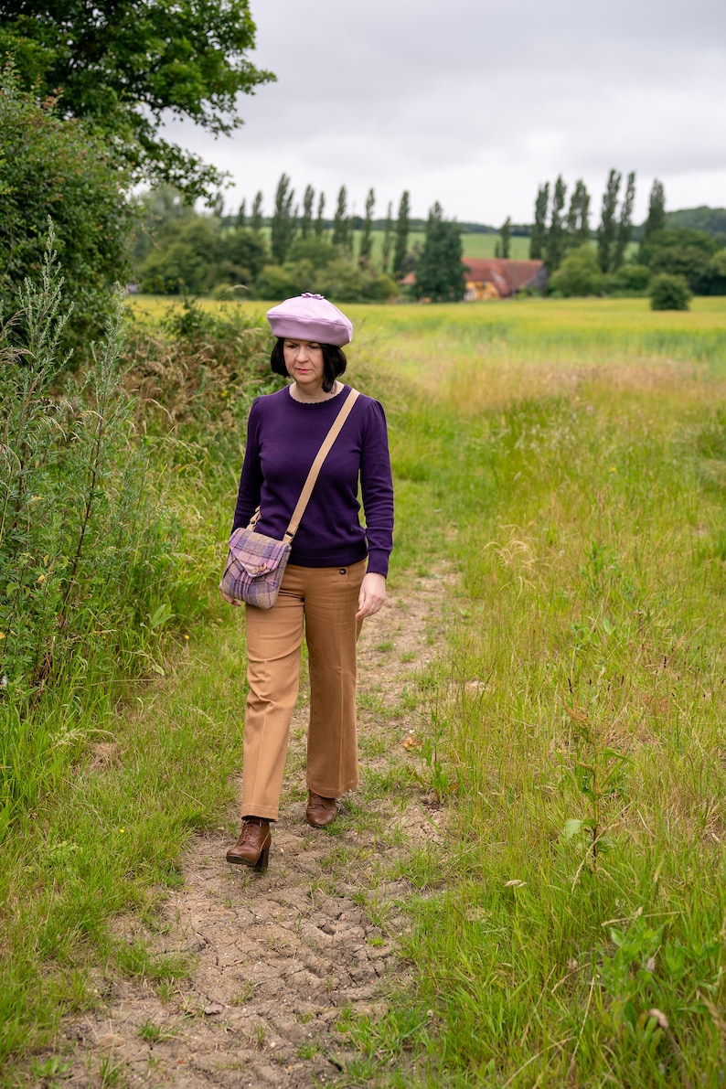 Pu&ograve; includere: Una donna con un maglione viola, pantaloni marroni e un berretto viola chiaro cammina su un sentiero di terra in un campo. Porta una borsa a tracolla a quadri.