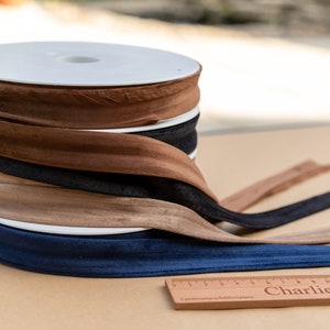 May include: A stack of five spools of brown, beige, black, and blue fabric trim. The spools are on a brown surface with a wooden ruler that says "Charlie's Aunt Sue" in front of them.