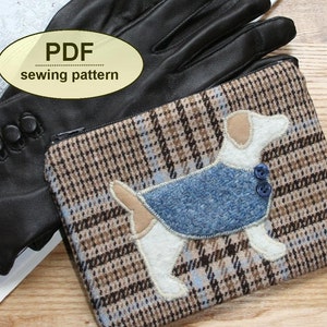 Könnte beinhalten: Eine braune und weiße karierte Reißverschlusstasche mit einem weißen Hund, der eine blaue Weste und zwei blaue Knöpfe trägt. Die Tasche liegt neben einem schwarzen Lederhandschuh mit Knöpfen.