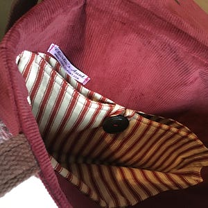 K&ouml;nnte beinhalten: Burgunderfarbene Cord-Tragetasche mit gestreifter Innentasche und Knopfverschluss. Die Tasche hat einen gewebten Griff und ein Etikett mit der Aufschrift "Charlie's Aunt".