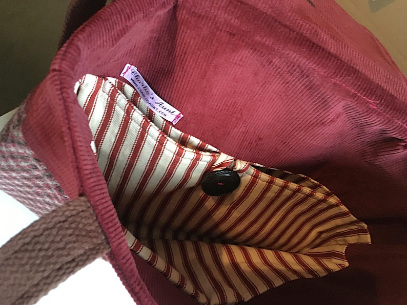 K&ouml;nnte beinhalten: Burgunderfarbene Cord-Tragetasche mit gestreifter Innentasche und Knopfverschluss. Die Tasche hat einen gewebten Griff und ein Etikett mit der Aufschrift "Charlie's Aunt".