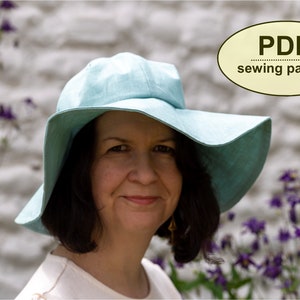 May include: A woman wearing a light blue sun hat with a wide brim. The hat is made of a soft, lightweight fabric. The woman is smiling and looking at the camera. The background is a white wall with purple flowers.