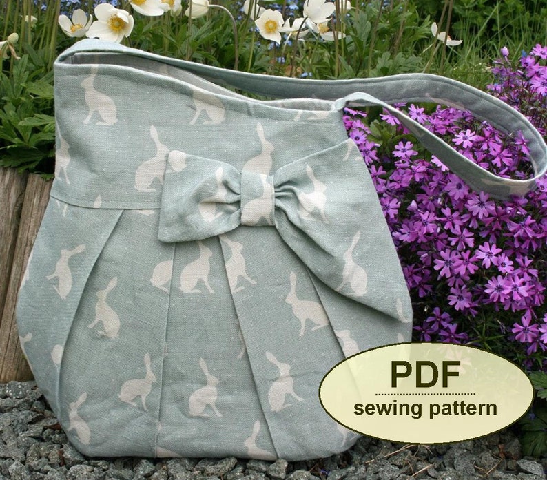May include: A light blue fabric tote bag with a white bunny rabbit print and a large bow. The bag has a pleated design and a long strap. The text "PDF sewing pattern" is printed on a white oval label.