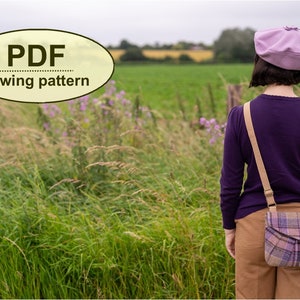 Pu&ograve; includere: Una donna con un maglione viola, pantaloni marroni e un berretto lavanda &egrave; in piedi in un campo. Indossa una borsa a tracolla a quadri con una tracolla marrone. Il testo "PDF sewing pattern" &egrave; in un ovale giallo nell'angolo in alto a sinistra dell'immagine.