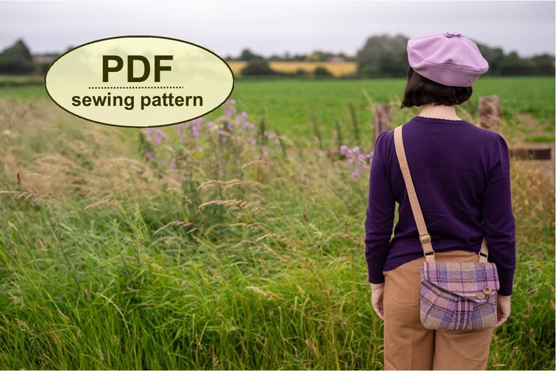 Pu&ograve; includere: Una donna con un maglione viola, pantaloni marroni e un berretto lavanda &egrave; in piedi in un campo. Indossa una borsa a tracolla a quadri con una tracolla marrone. Il testo "PDF sewing pattern" &egrave; in un ovale giallo nell'angolo in alto a sinistra dell'immagine.