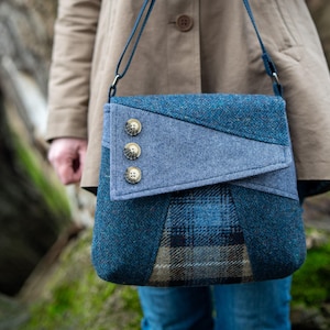 Op de afbeelding: Blauwe tweed crossbodytas met een geometrisch ontwerp. De tas heeft een klepsluiting met drie decoratieve knopen. De tas heeft een blauwe band en is gemaakt van verschillende blauwe en grijze stoffen.