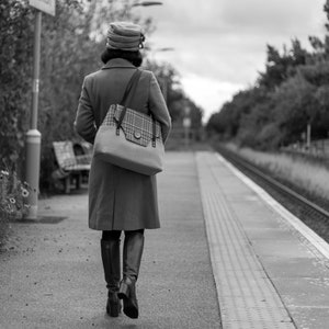 May include: A woman in a brown coat and a hat walks away from the camera on a train platform. She is carrying a plaid tote bag.