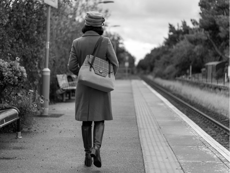 May include: A woman in a brown coat and a hat walks away from the camera on a train platform. She is carrying a plaid tote bag.