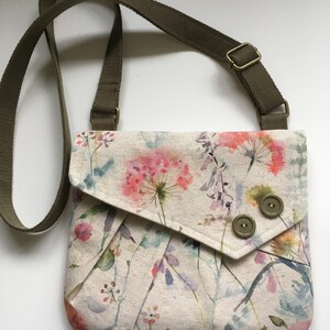 Pu&ograve; includere: Una piccola borsa a tracolla con motivo floreale, tracolla verde e due bottoni marroni. La borsa ha una chiusura a lembo e uno sfondo crema con fiori in stile acquerello in tonalit&agrave; rosa, viola e verde.