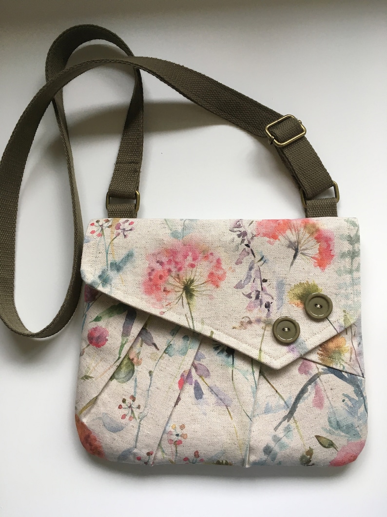Pu&ograve; includere: Una piccola borsa a tracolla con motivo floreale, tracolla verde e due bottoni marroni. La borsa ha una chiusura a lembo e uno sfondo crema con fiori in stile acquerello in tonalit&agrave; rosa, viola e verde.