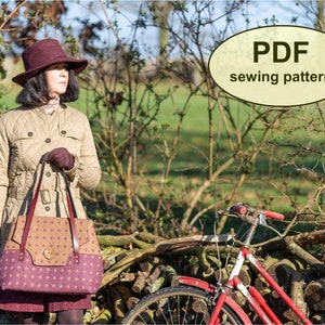 May include: A woman wearing a brown quilted jacket and a burgundy hat is holding a brown and purple tote bag with a large button detail. The bag has a brown leather strap. The woman is standing in front of a pile of logs and a red bicycle.