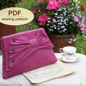May include: A pink fabric clutch with a large bow and three buttons. The clutch is sitting on a piece of sheet music on a white tablecloth. The text "PDF sewing pattern" is visible in the image.
