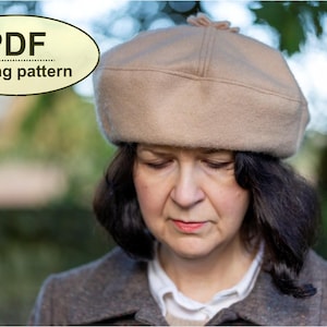 Könnte beinhalten: Eine Frau trägt eine beige Baskenmütze mit einem kleinen Bommel oben. Sie trägt eine braune Jacke und hat dunkles Haar.