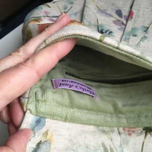 Pu&ograve; includere: Una borsa in tessuto di velluto a coste verde con una tasca con cerniera e un'etichetta viola che recita "Frou Frou & Aunt" e "Handmade Artisan", con una stampa floreale all'esterno.