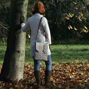 May include: A woman wearing a brown hat and a white sweater is carrying a plaid shoulder bag with a brown leather strap. The bag has a flap closure with a metal buckle and a decorative button.