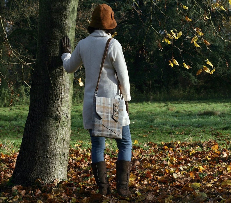 May include: A woman wearing a brown hat and a white sweater is carrying a plaid shoulder bag with a brown leather strap. The bag has a flap closure with a metal buckle and a decorative button.