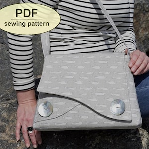 Può includere: Borsa a tracolla grigio chiaro con motivo a pesce e due grandi bottoni rotondi e iridescenti. La borsa ha una chiusura con patta e una lunga tracolla. Il testo "PDF sewing pattern" è in un ovale giallo in alto a sinistra.
