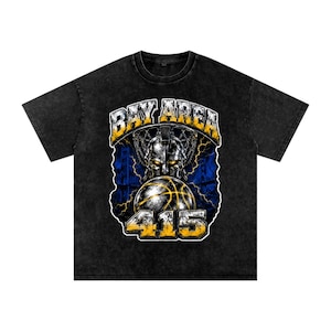 Op de afbeelding: Zwart T-shirt met een grafisch ontwerp met een basketbal, een gladiatorenhelm en de woorden "BAY AREA" en "415" in geel en wit. Het ontwerp is geplaatst tegen een blauwe en gele bliksemachtergrond.