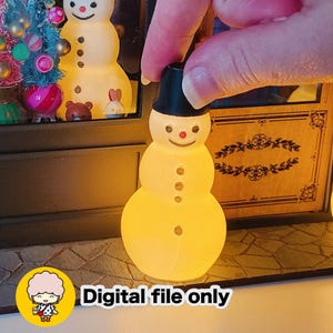 Puede incluir: Una figura de muñeco de nieve brillante con un sombrero de copa negro, una nariz roja y tres botones negros. El muñeco de nieve está iluminado con una cálida luz amarilla. También incluye el texto "Digital file only".