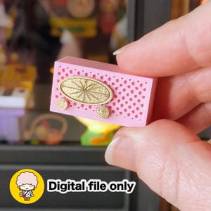 Puede incluir: Una radio miniatura rectangular de color rosa con una rejilla de altavoz perforada, un detalle central ovalado de color dorado y dos pequeños botones circulares dorados en la parte frontal. La imagen incluye un icono amarillo con un personaje y el texto Digital file only.