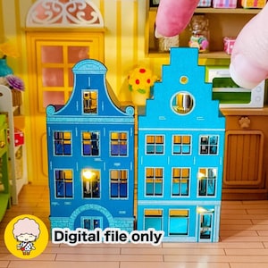 May include: Two miniature blue houses with illuminated windows, featuring a Dutch architectural style. The houses are made of paper or cardboard and are part of a dollhouse or craft project. The text "Digital file only" is visible.