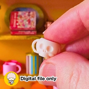 May include: A miniature white skull-shaped mug with a small handle. The object is held between fingers. The image includes a yellow circular icon featuring a cartoon character and the black text Digital file only on a white background.