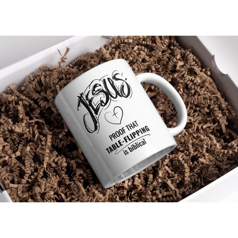 Puede incluir: Taza de cer&aacute;mica blanca con la palabra "JESUS" en fuente negra estilizada, un coraz&oacute;n y la frase "PROOF THAT TABLE-FLIPPING is biblical." La taza est&aacute; dentro de una caja blanca llena de material de embalaje marr&oacute;n.