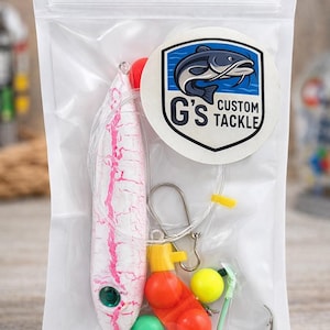 May include: A clear plastic bag containing fishing tackle. The bag includes a white and pink lure, various colourful floats, a green hook, and a sticker with a fish graphic and the text "G's Custom Tackle".
