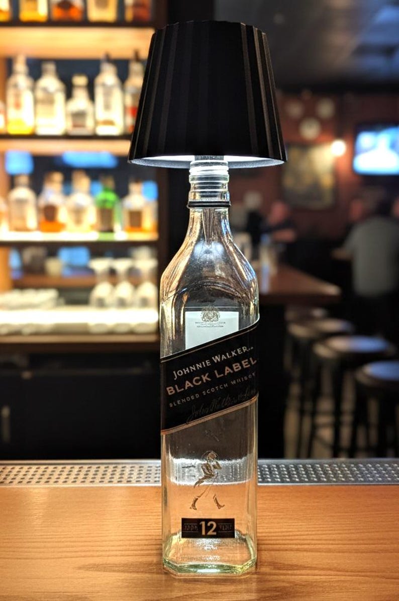 May include: A bespoke lamp crafted from a Johnnie Walker Black Label whisky bottle. It has a black lampshade and a clear glass bottle base. The label displays "Johnnie Walker Black Label Blended Scotch Whisky".