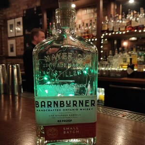 May include: A clear glass bottle of "Barnburner" handcrafted Ontario whisky, with a cork stopper. The label includes text such as "Maverick Distillery" and "Small Batch". The bottle is lit from within with green lights, and sits on a wooden bar.