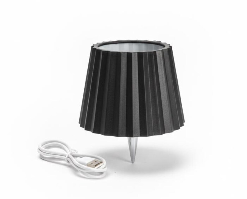 May include: A black, pleated lampshade-style light fixture with a silver base and a white USB charging cable. The lamp has a conical shape with vertical pleats. The cable is coiled next to the lamp.