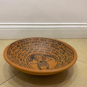 May include: A round, earthenware bowl with a light brown hue. The bowl features black text around the rim and a central illustration of a figure. The text appears to be in a script, possibly Hebrew. The bowl is on a light-colored surface.