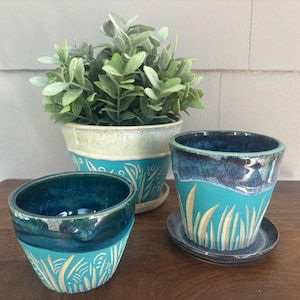 May include: Three ceramic planters in shades of teal and blue, with a cream-colored accent. The planters feature a hand-painted design of tall grass. One planter has a matching saucer. A green plant sits in the largest planter.