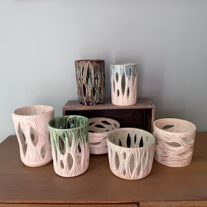 May include: A collection of six ceramic vessels featuring cut-out patterns resembling tree bark or organic textures. The pieces vary in height and color, including shades of pale pink, green, and dark brown, with some displaying a gradient glaze finish.