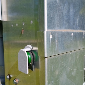May include: A metallic industrial control panel featuring a brushed steel surface with visible rivets and a green circular push button protected by a gray plastic guard. The unit has a rectangular structure with sharp edges and a small insect on the side.