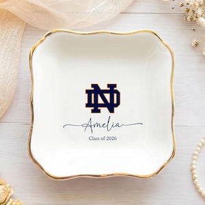 Puede incluir: Un plato cuadrado de cerámica blanca con bordes festoneados y un borde pintado en oro. El centro presenta un logotipo de monograma azul oscuro de Notre Dame sobre el nombre Amelia en letra cursiva y el texto Class of 2026 en fuente simple.