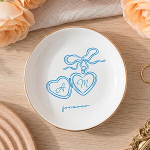 May include: A round white ceramic trinket dish with a gold-toned rim. The center features a blue line-art illustration of two heart-shaped lockets tied with a bow, with the letters A and M inside the hearts and the word forever written below.
