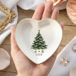 Könnte beinhalten: Eine weiße, herzförmige Keramikschale mit goldfarbenem Rand. In der Mitte befindet sich eine Grafik eines geschmückten Weihnachtsbaums mit einem goldenen Stern an der Spitze, den Kleinbuchstaben c + b und der Jahreszahl 2026 in Schwarz.