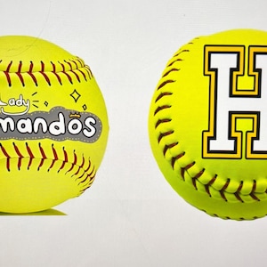 May include: Two yellow softballs. One features the text "Lady Commandos" in gray and white, with a crown and diamond accents. The other displays a large white "H" with a black and yellow outline.