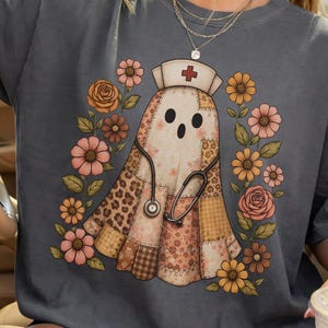 Puede incluir: Una camiseta gris carbón de manga corta con un gráfico central de un fantasma vestido de enfermero con estetoscopio, que lleva un gorro de enfermería con una cruz roja, rodeado por un patrón floral de flores marrones, naranjas y rosas.